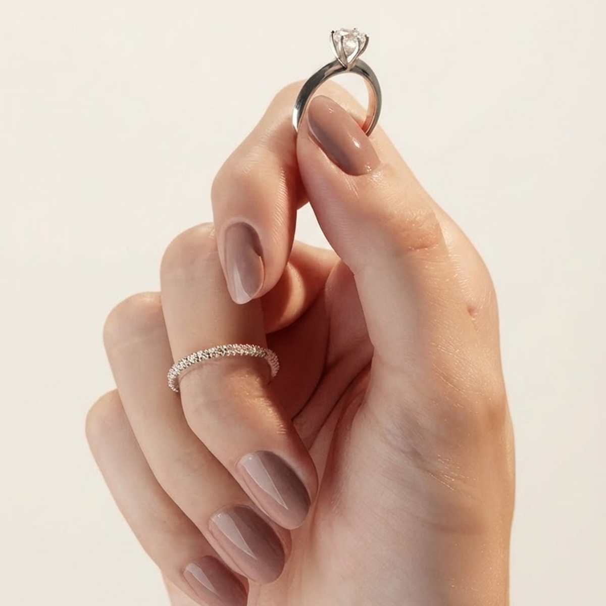 Hand holding up a solitaire engagement ring with a six-prong setting, a pavé band worn on another finger, on a cream background