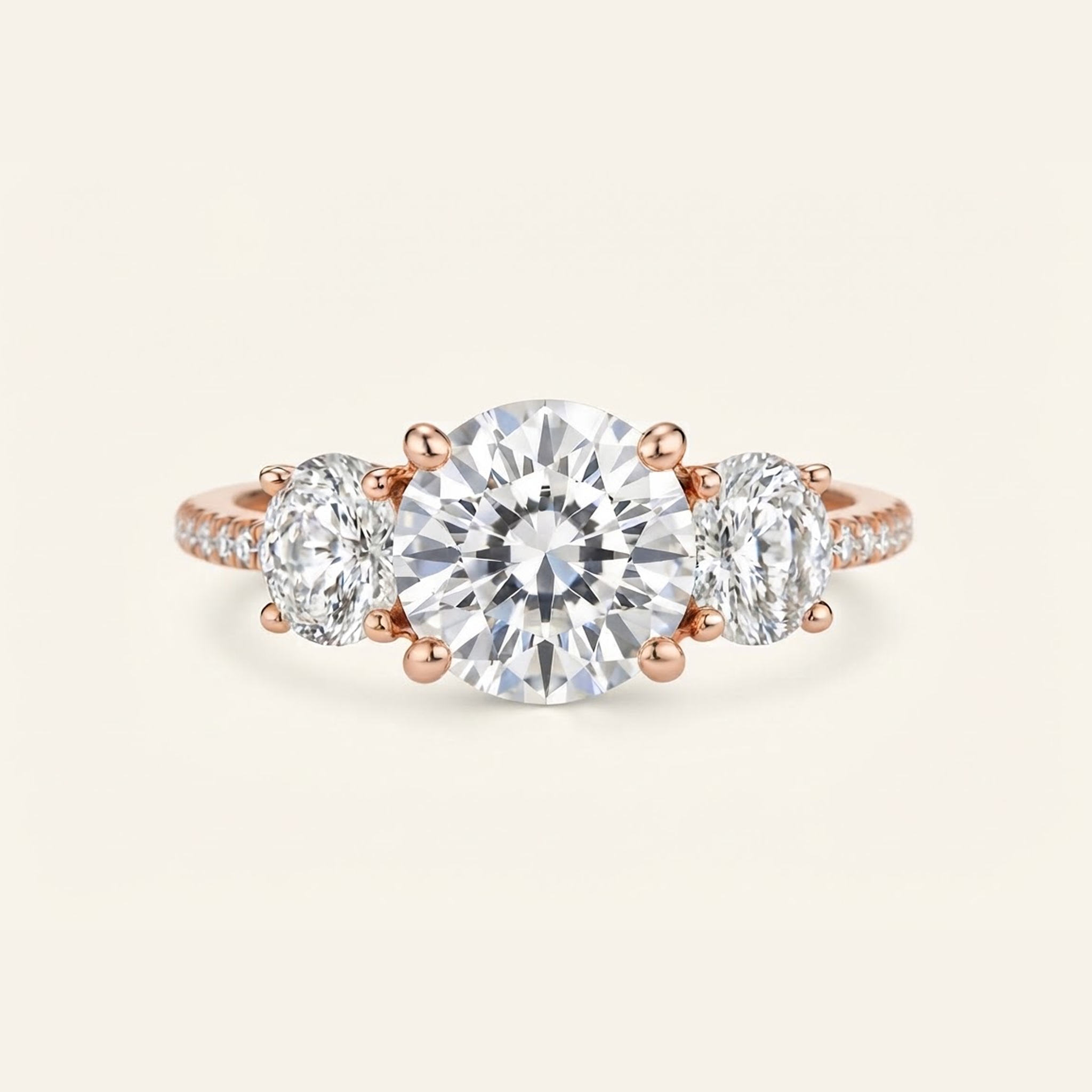Celia in rose gold, a three-stone engagement ring featuring a large round center diamond flanked by two oval side diamonds, with small pavé accents on the band, set against a solid cream background.

variant: rose-gold