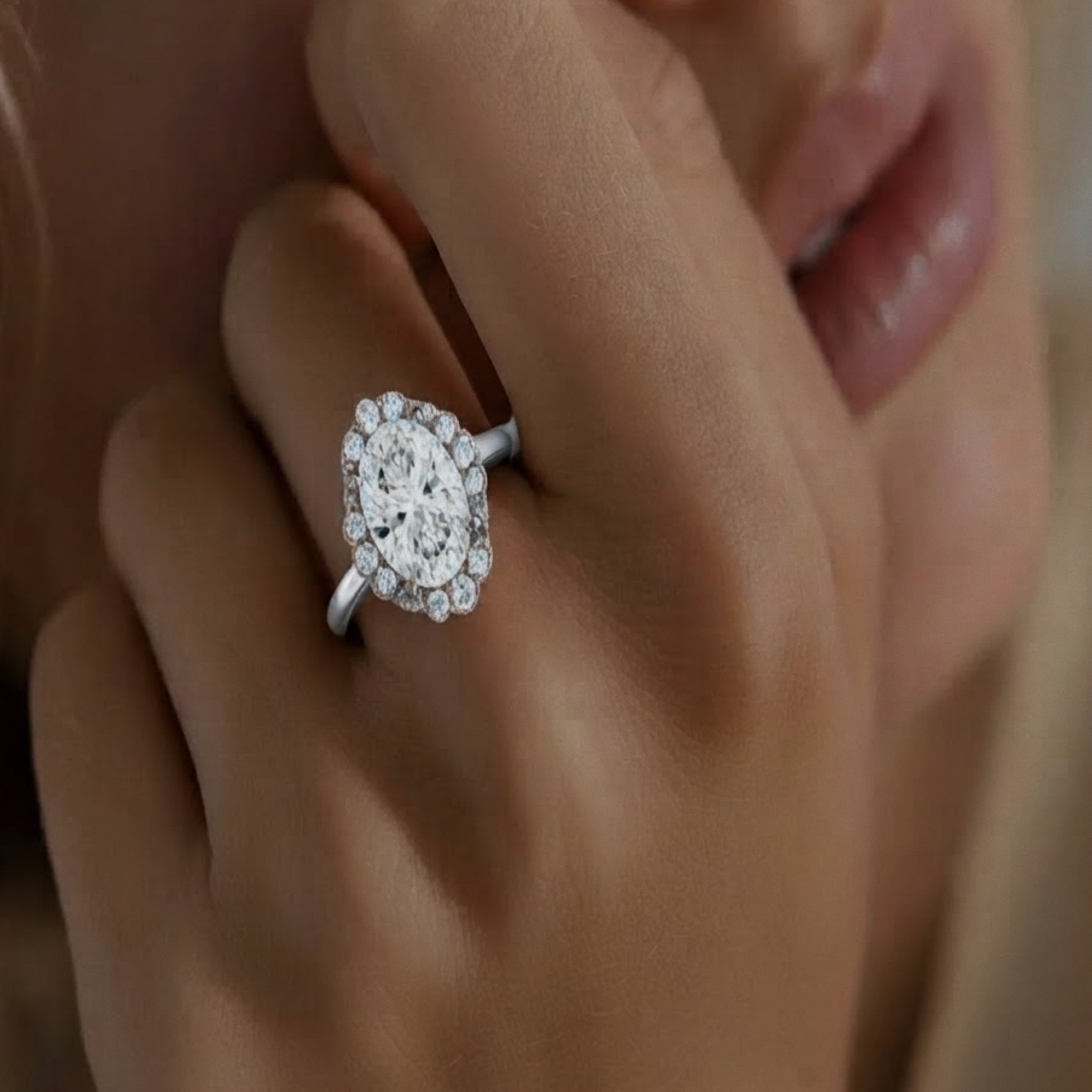 Elise Ring in white gold, an oval halo engagement ring, worn on a hand held close to the face.

variant: white-gold