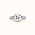 Noelle engagement ring in white gold with a round diamond in four claw prongs and a pavé diamond band, on a cream background
variant: white-gold