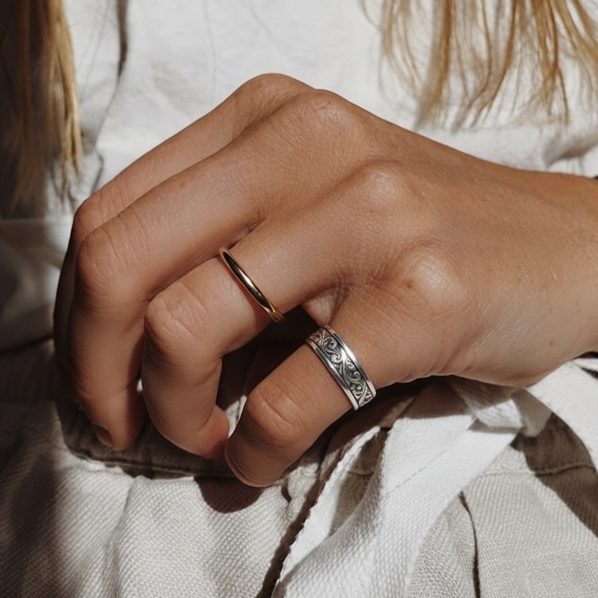 A close-up of a person's hand wearing including Verona ring in silver, a textured, patterned band ring and a simple thin band ring, set against a soft white fabric background.

variant: silver