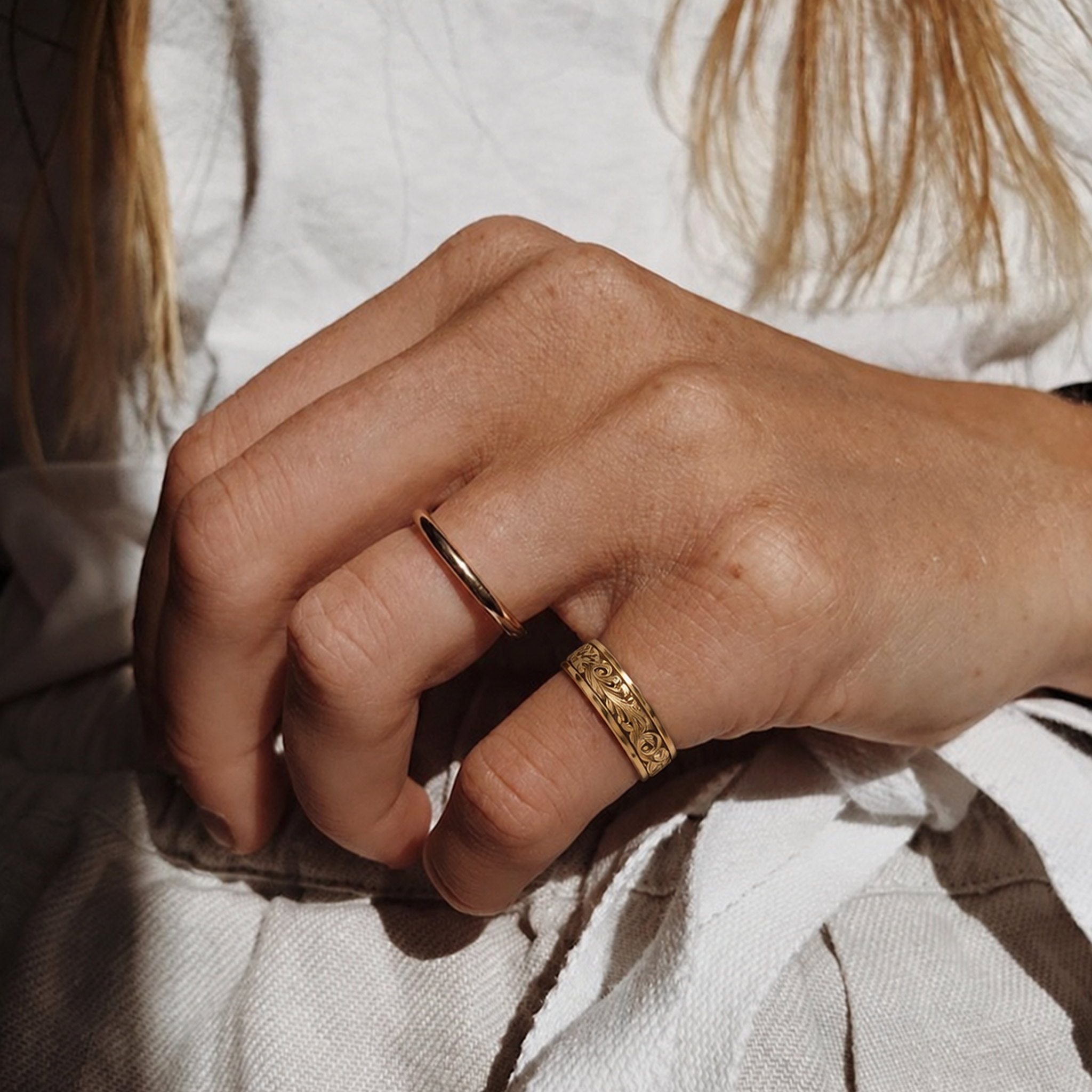 A close-up of a person's hand wearing two gold rings, including Verona ring, a textured, patterned band ring and a simple thin band ring, set against a soft white fabric background.

variant: yellow-gold