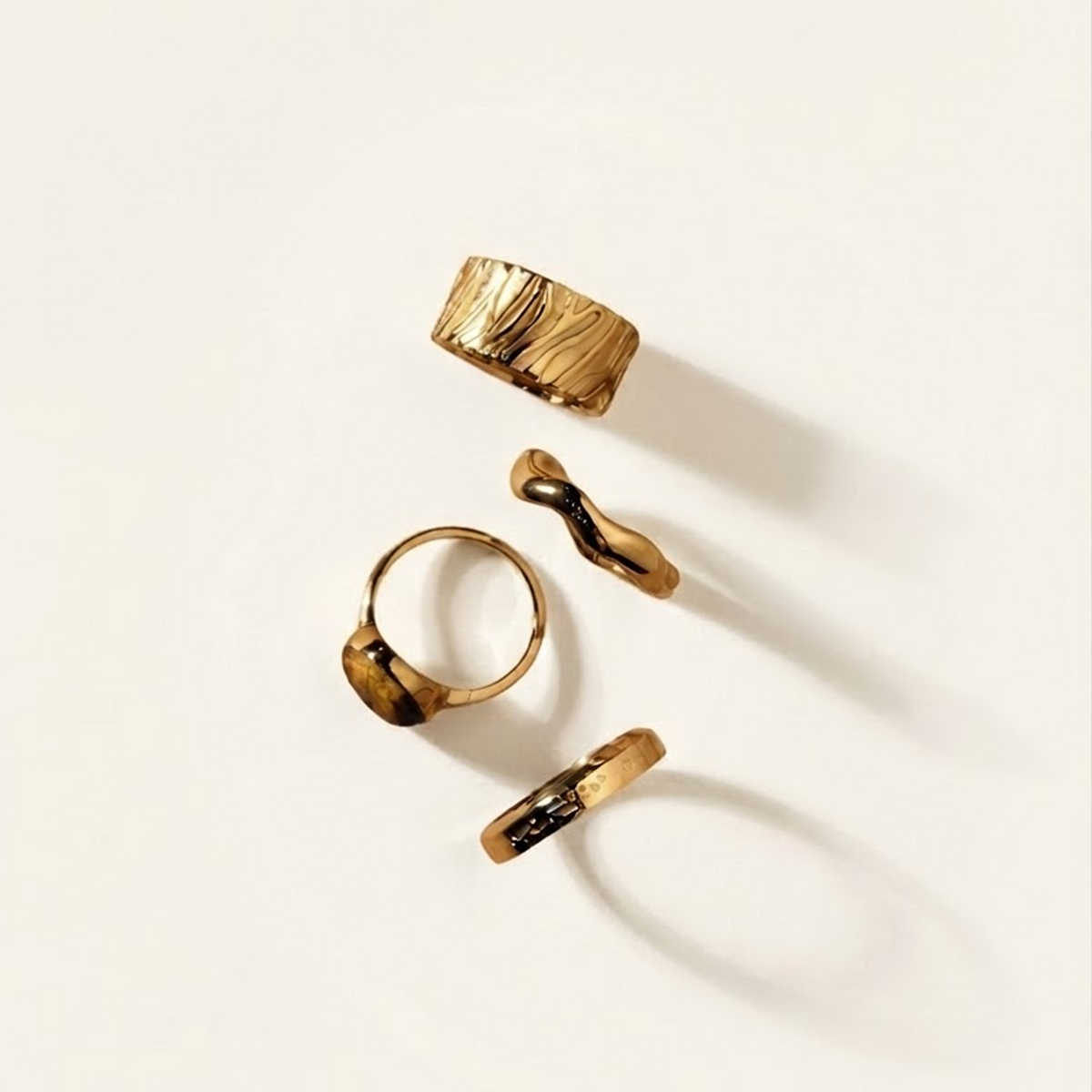 Four gold rings with organic, wavy, and textured designs arranged on a clean off-white background with soft shadows.