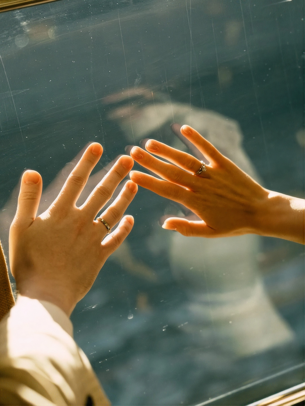 Two hands pressed against a sunlit window, each wearing a gold ring, with a couple's reflection in the glass.