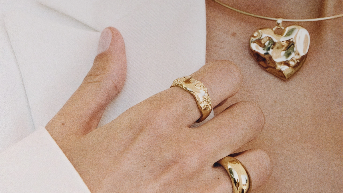 Close-up of a person wearing gold jewelry, including a textured heart-shaped pendant necklace and two rings on the fingers, with a white blazer visible.