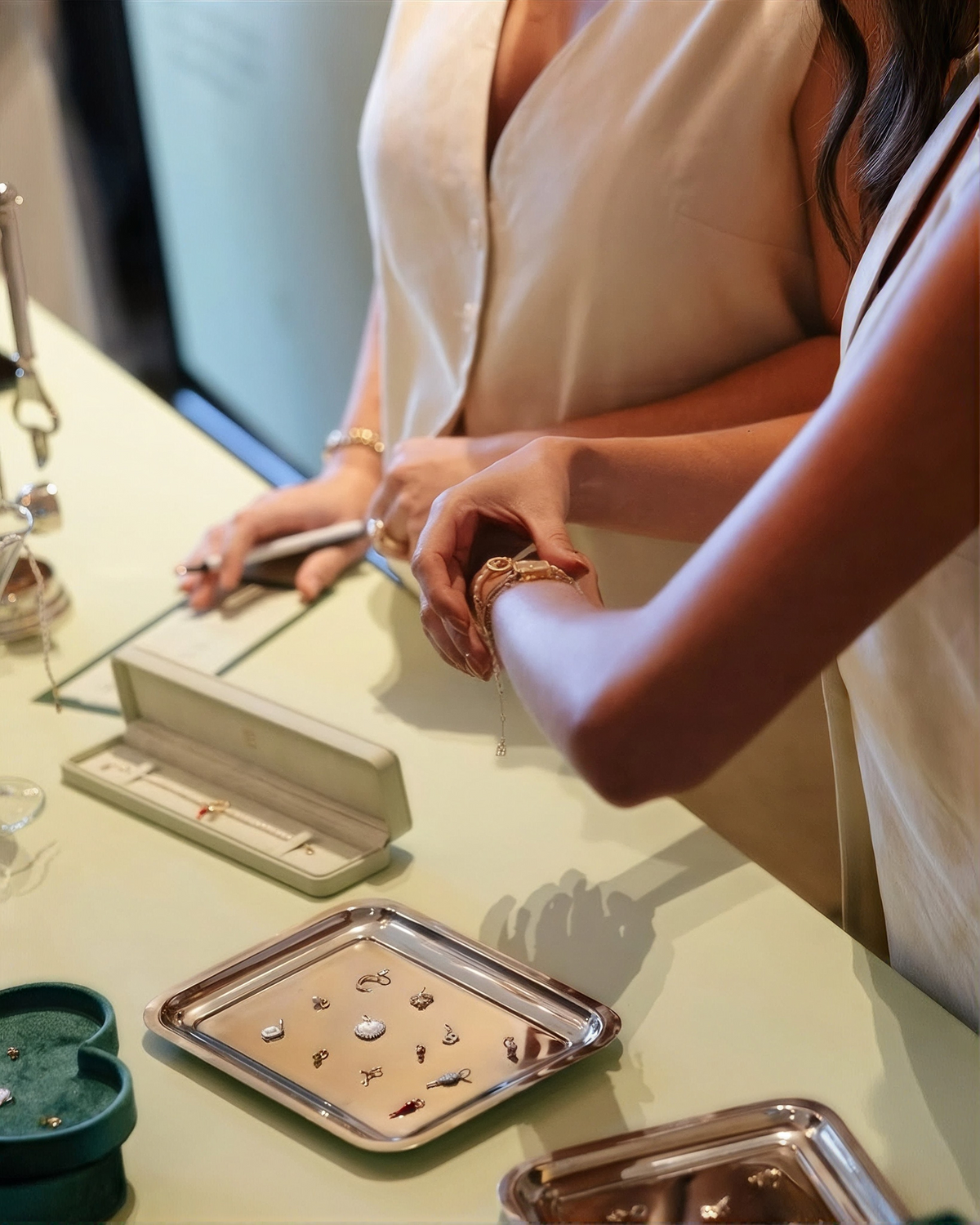 A close-up lifestyle shot of a jewelry display counter where a model in a cream sleeveless top is shown wearing a delicate gold chain bracelet. Several silver trays containing various small jewelry pieces are arranged on the light-colored counter next to open jewelry boxes.