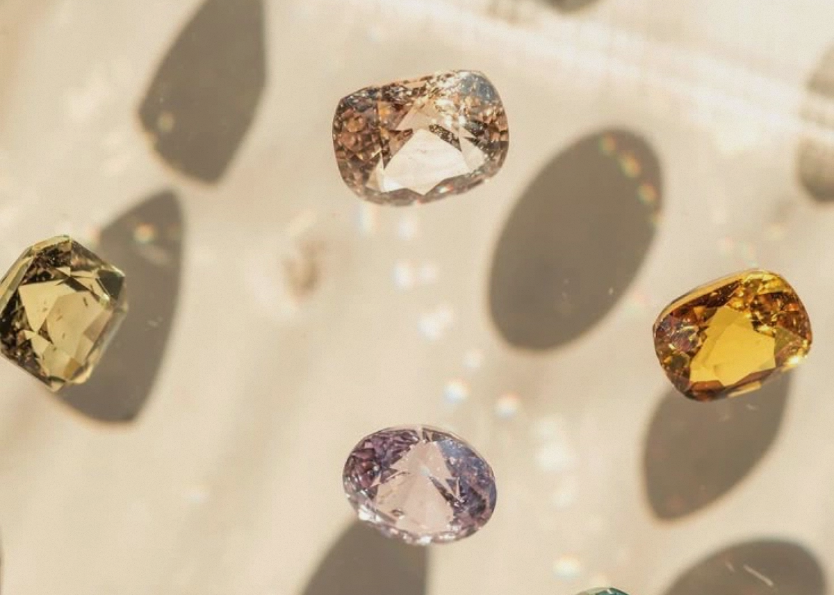 Several loose oval-cut gemstones in different colors, including yellow, purple, blue-green, and clear, arranged on a glossy light surface with strong reflections and shadows.