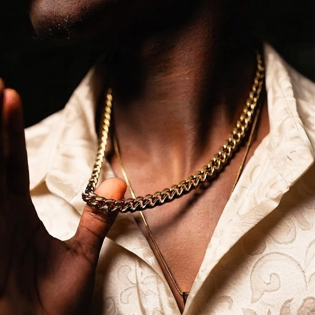 A close-up lifestyle shot of a person wearing a thick gold Cuban link chain necklace over a cream-colored patterned shirt against a dark background.