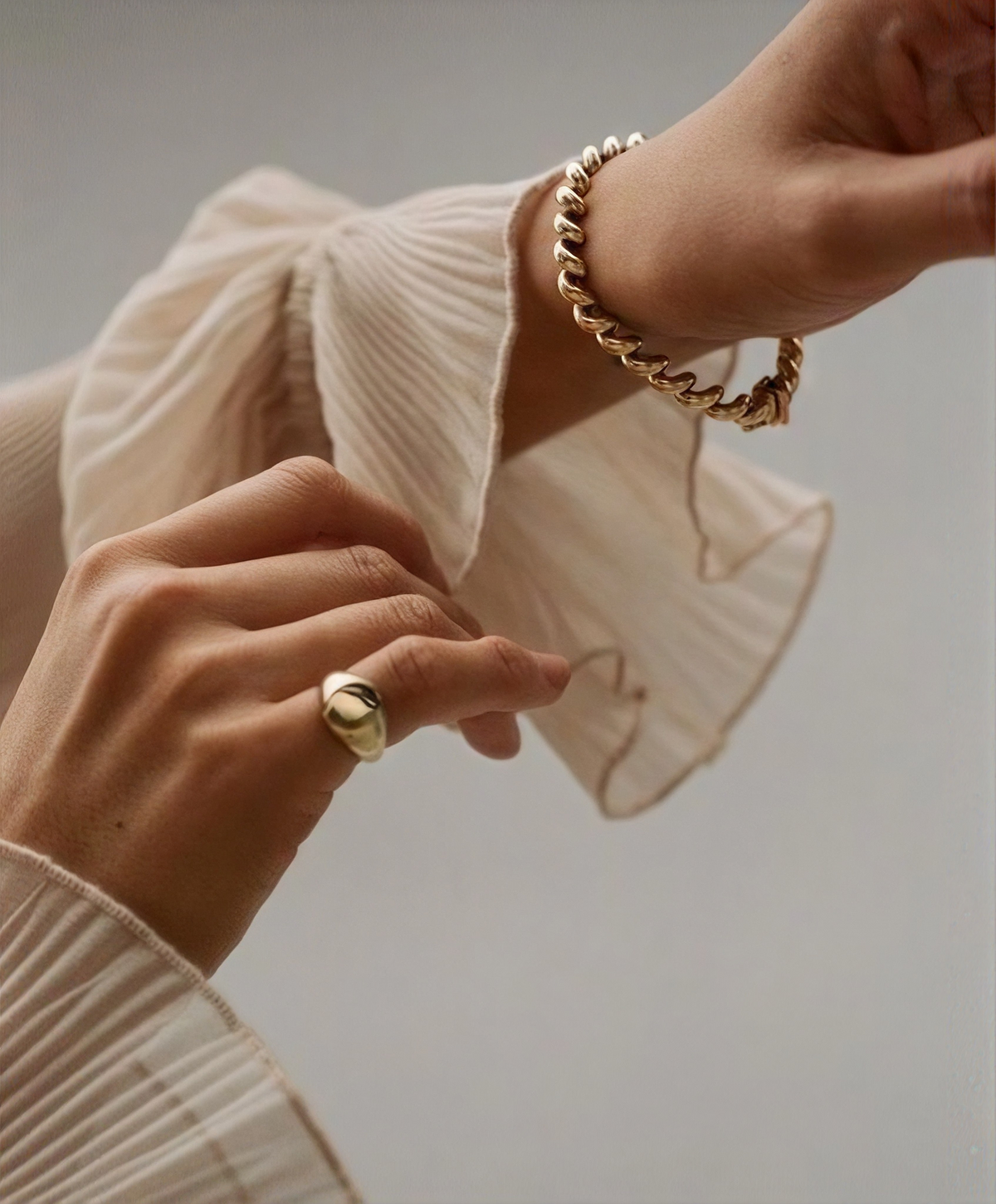 A close-up lifestyle shot of a person wearing a textured gold ring on one finger and a chunky gold chain bracelet on the wrist, paired with a pleated cream-colored blouse against a soft gray background.