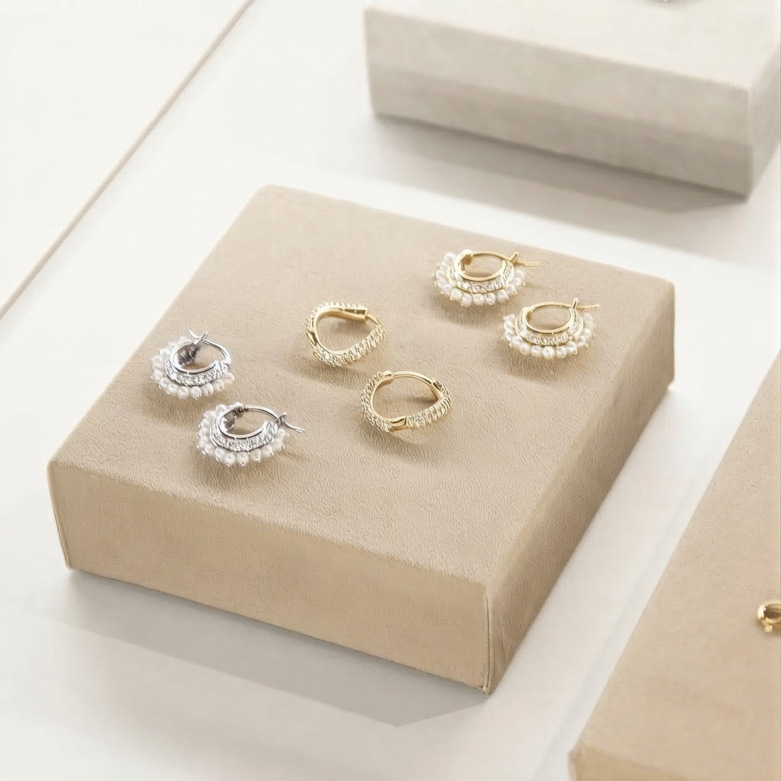 Four small hoop earrings displayed on beige jewelry boxes against a clean white background. Two earrings are gold-toned with textured pavé detailing, and two are silver-toned with pearl-like bead detailing.