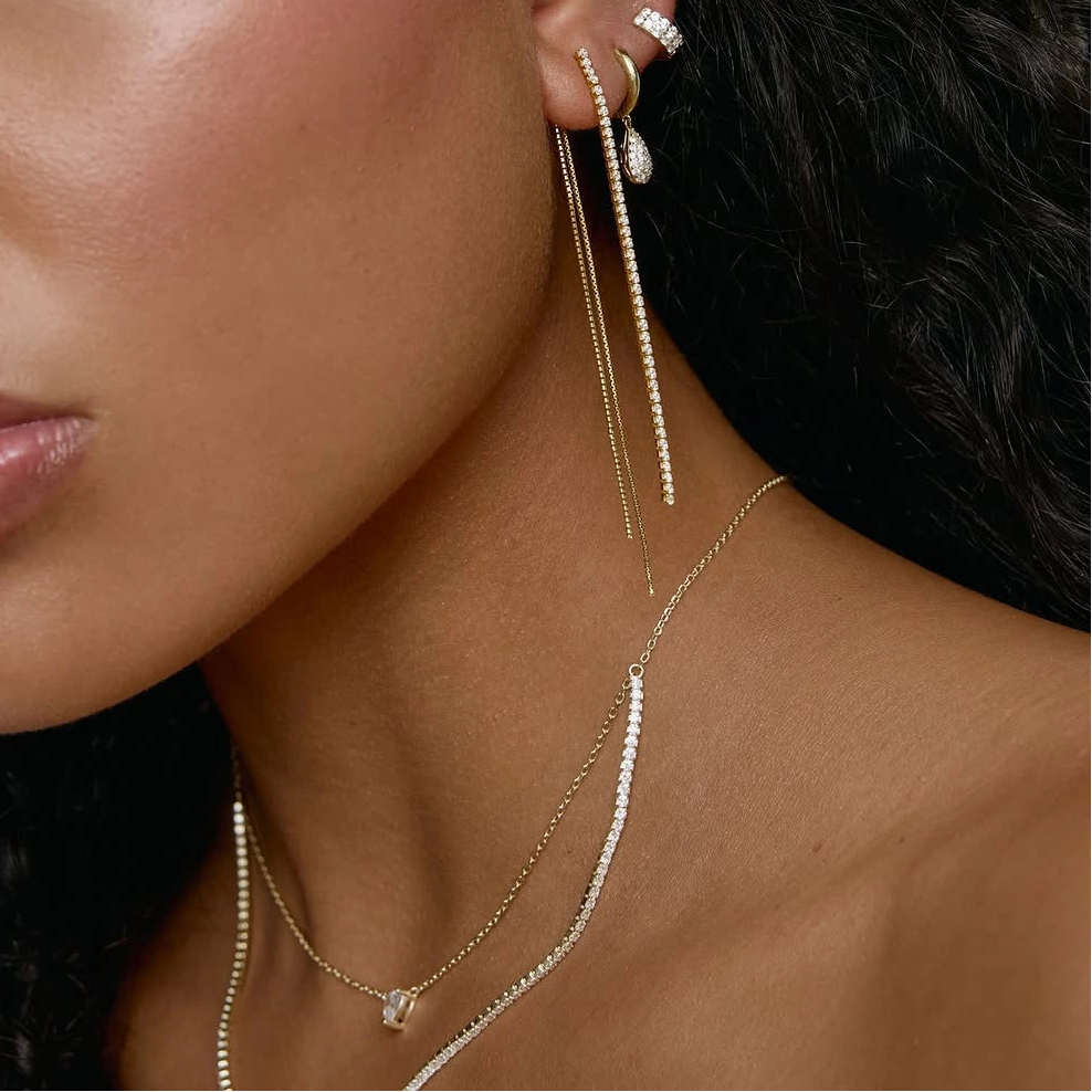 A close-up of a model wearing a pair of gold drop earrings with small clear stones and long dangling chains, along with delicate gold necklaces.
