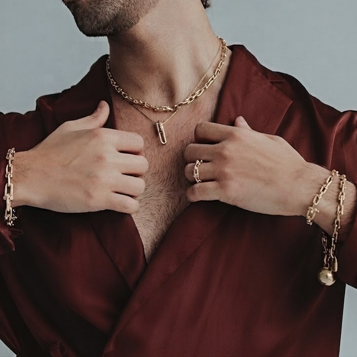 A man wearing a shiny burgundy satin shirt and gold chain jewelry, including a chain necklace, bracelet, and rings, posed against a solid light gray background.