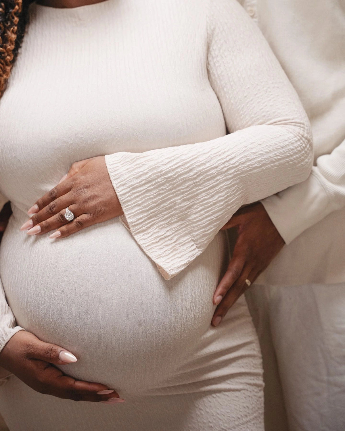 A close-up lifestyle shot of a pregnant woman wearing a textured cream-colored long-sleeve maternity dress, cradling her baby bump with another person's hand resting on her side.